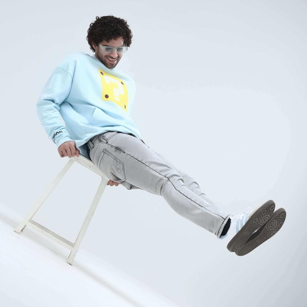Man sitting on a stool wearing a light blue sweatshirt and grey jeans, showcasing a casual and stylish outfit.
